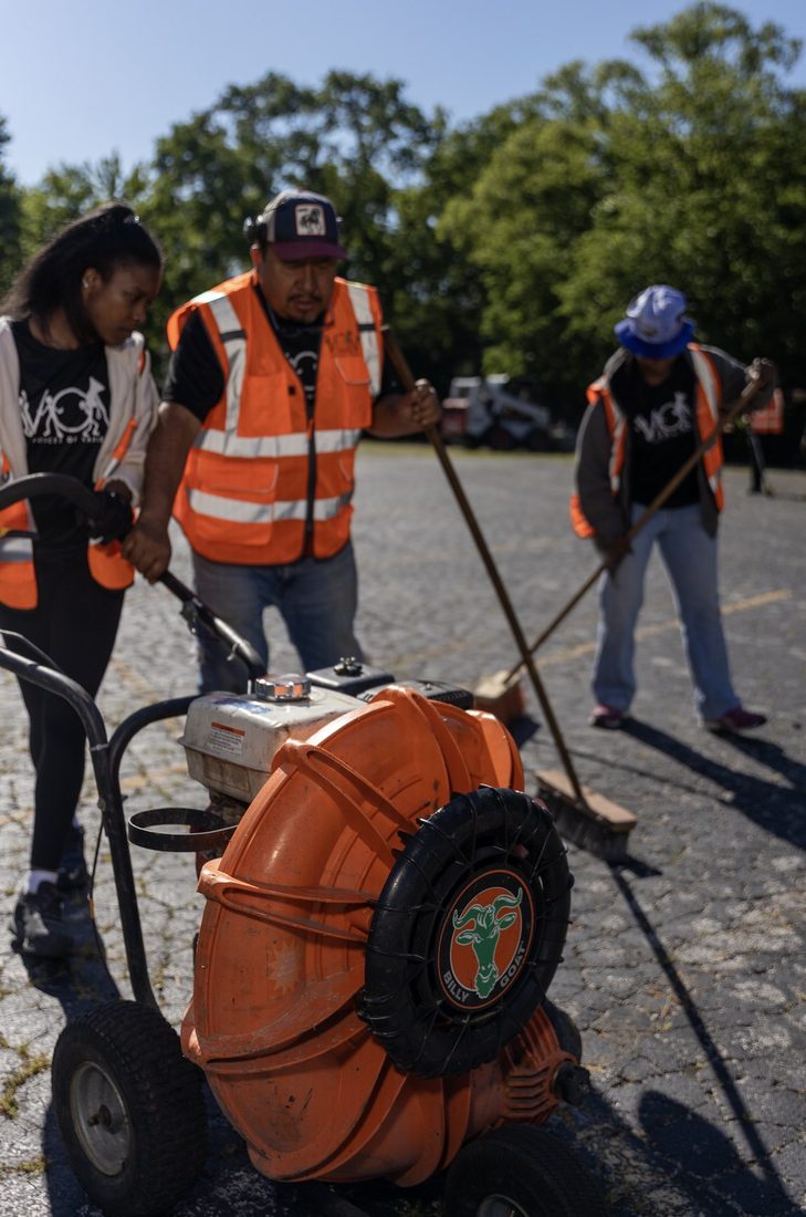 V.O.V crew in orange vests clearing a parking lot with a blower and brooms