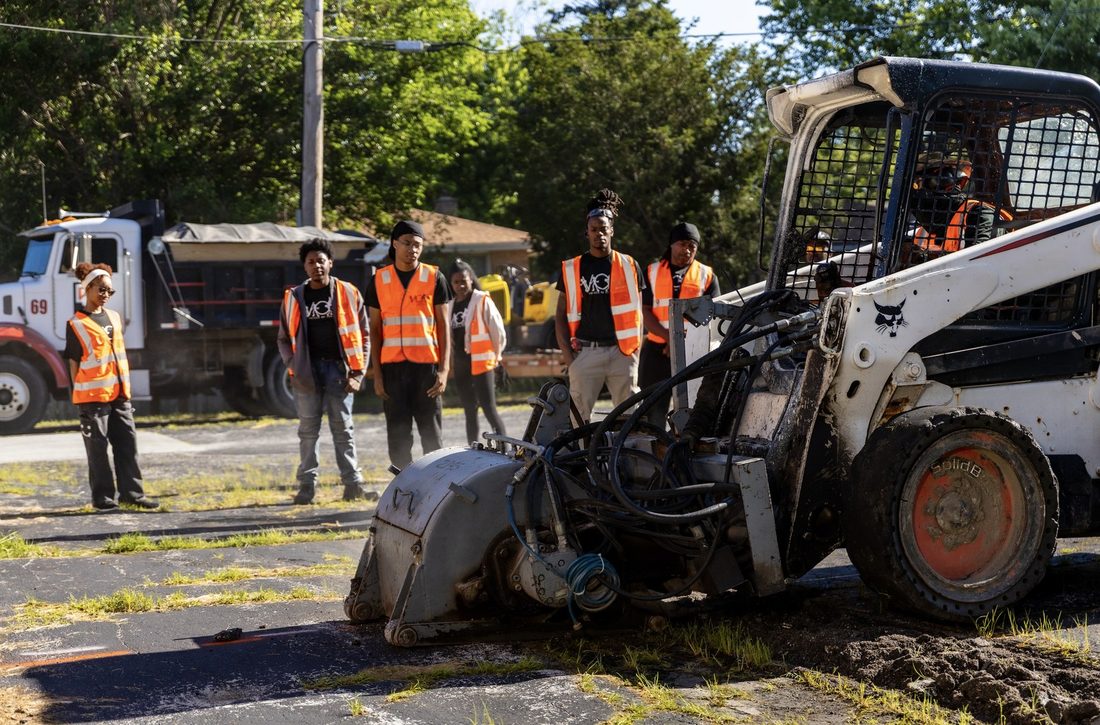 V.O.V work crew in safety vests watching a skid-steer saw cut pavement on a job site